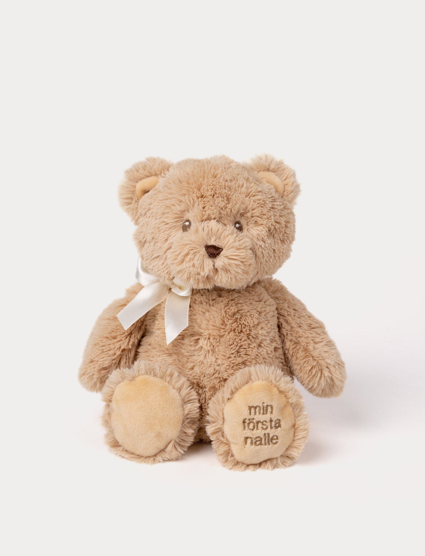 Soft beige teddy bear plush with a white ribbon and "min första nalle" embroidered on the paw.
